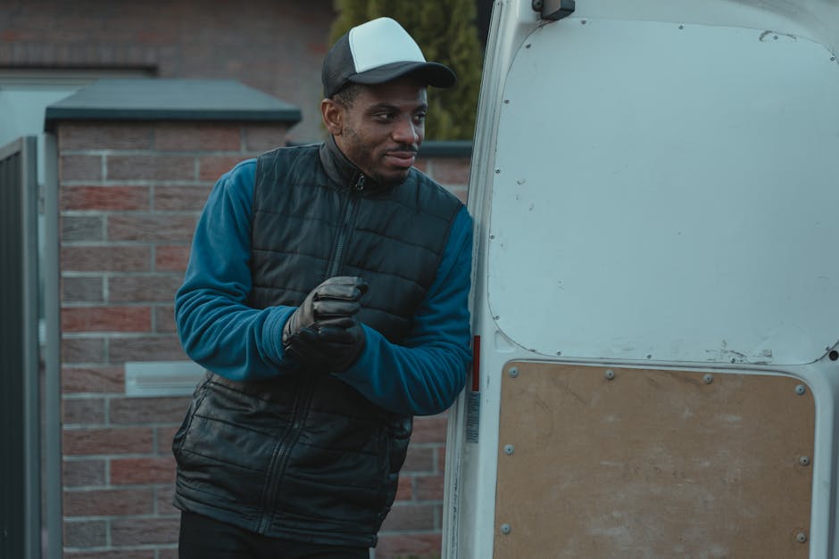 A man dressed in a dark padded vest over a blue long-sleeve shirt, wearing a grey and black baseball cap and black gloves, is standing outdoors next to a white van with an open rear door. He appears t